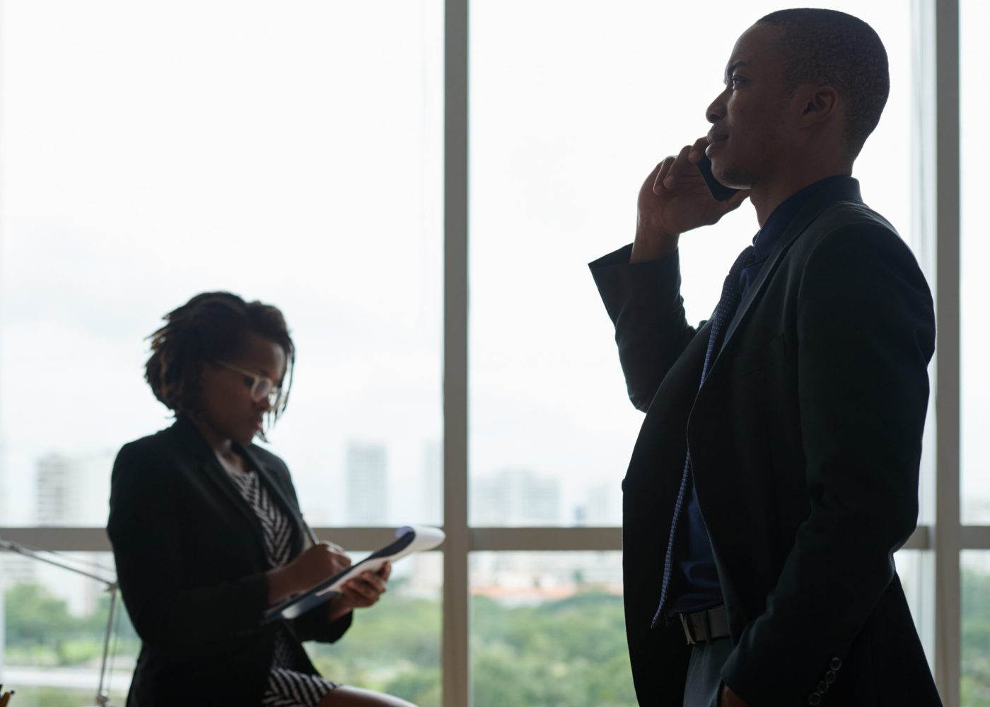 Young smiling businessman standing against big window and calling on the phone