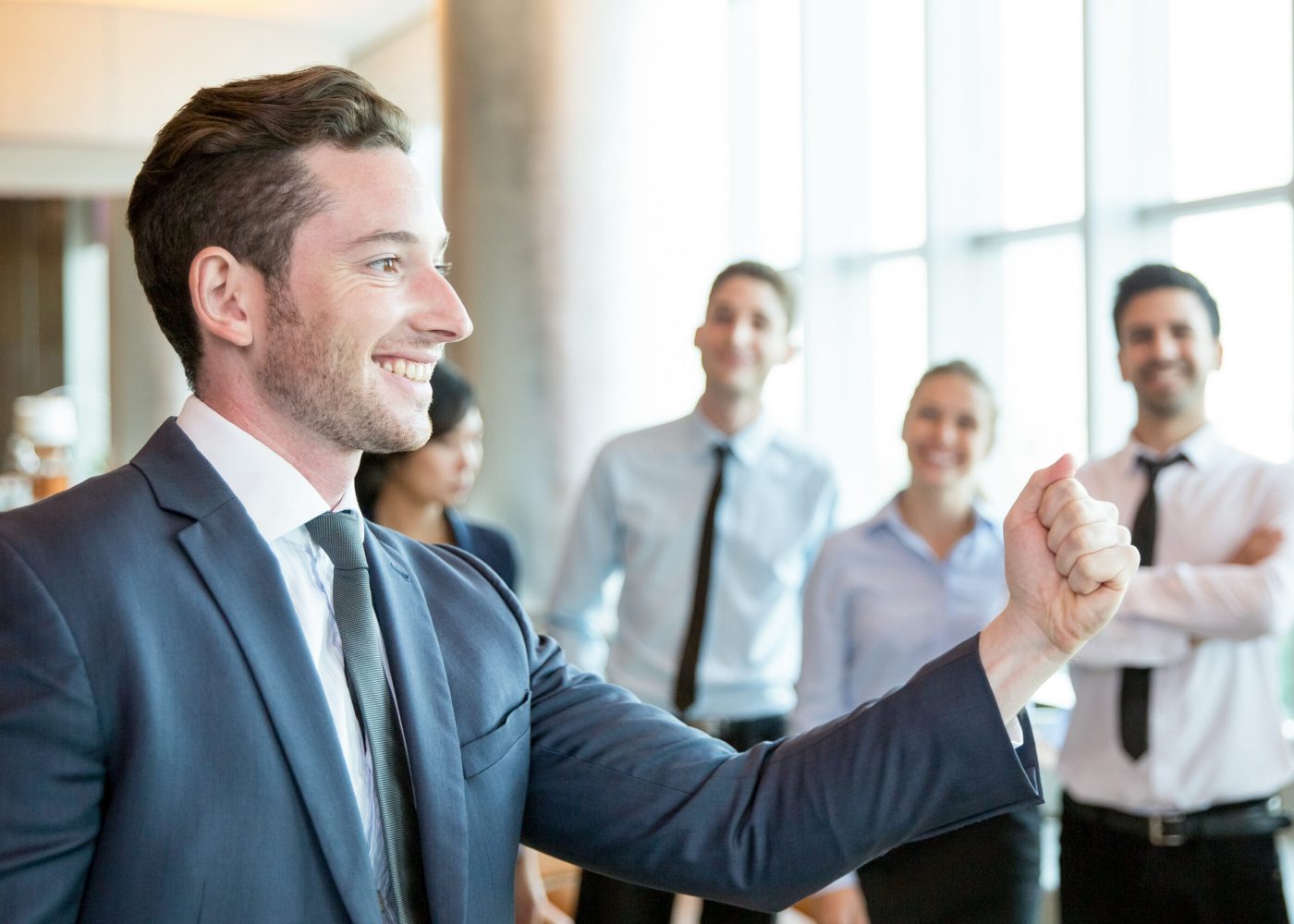 Cheerful leader motivating his business team. Handsome young politician telling his plan and showing fist as symbol of power. Positive business people standing in background. Strong company concept