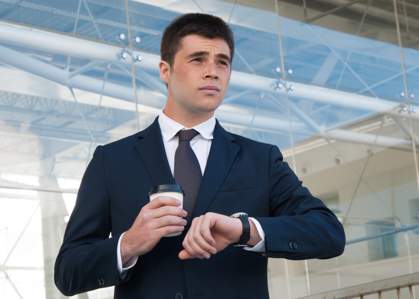 Pensive business man checking time on watch outdoors. Handsome guy holding plastic coffee cup and standing with building glass wall in background. Business appointment concept. Front view.