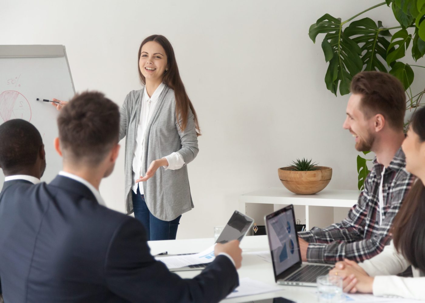 Smiling young employee or manager giving presentation working on flipchart in meeting room making business offer or reporting about project results, explaining new plan or idea to multi-ethnic team