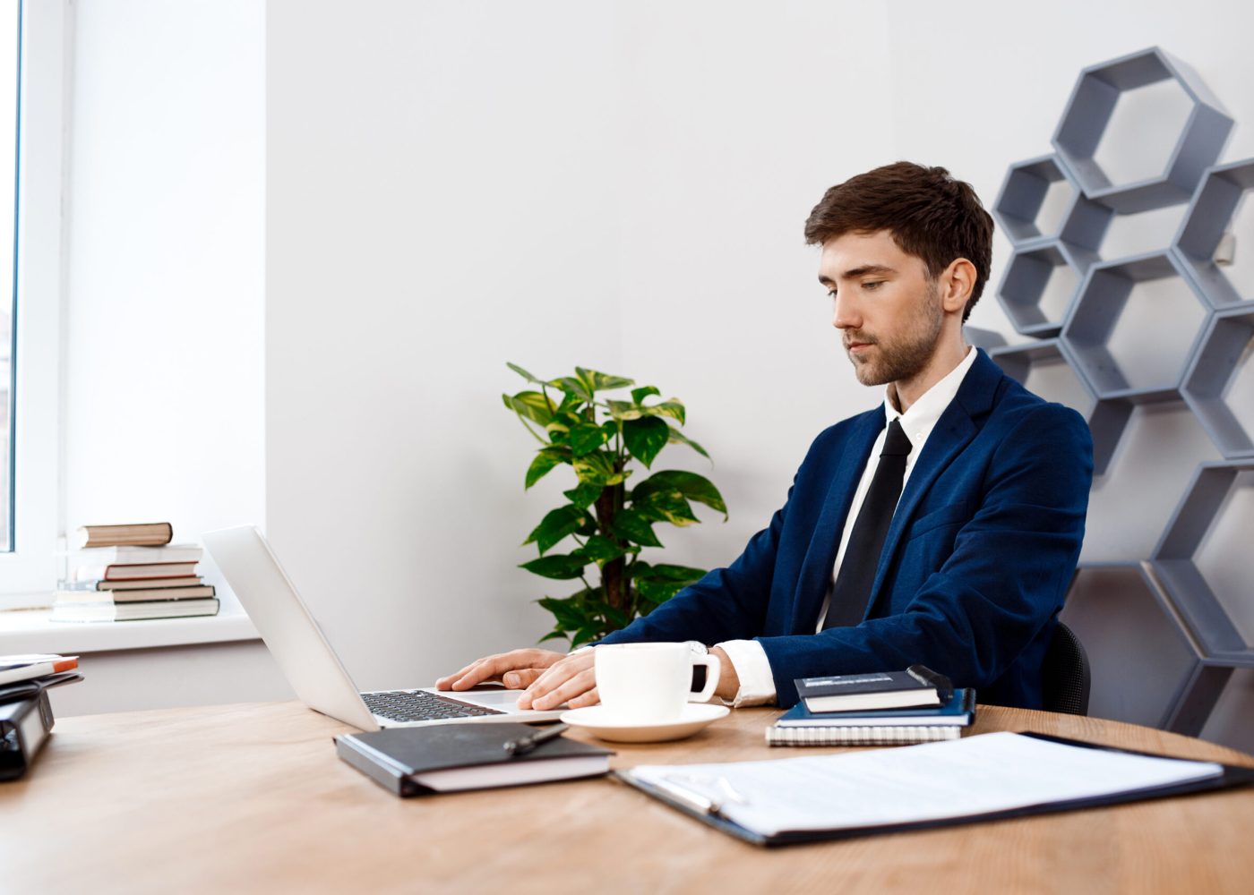 Young successful businessman in suit sitting at laptop at workplace, office background.