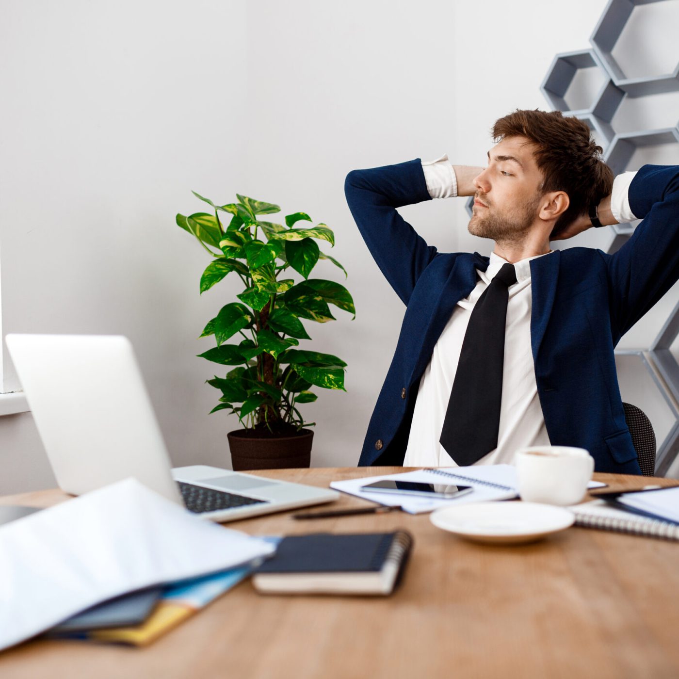 Young successful businessman in suit sitting at workplace, hands behind head, office background.